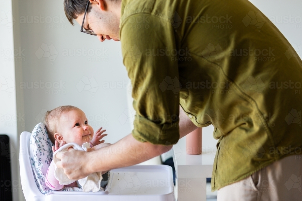 Father cleaning messy baby up after eating food in high chair - Australian Stock Image