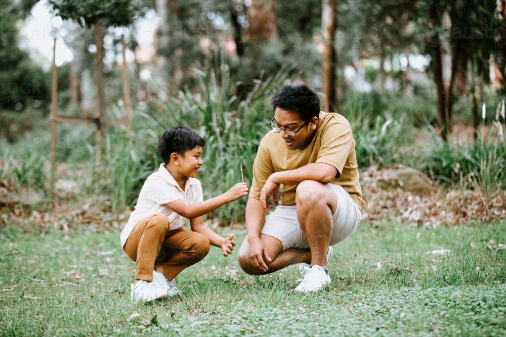 Father crouching on the grassy field of the park with son holding dry leaf - Australian Stock Image