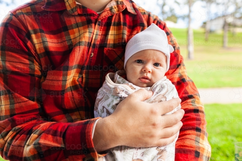 Father holding baby at park wrapped up in blanket and cap to keep warm in winter - Australian Stock Image