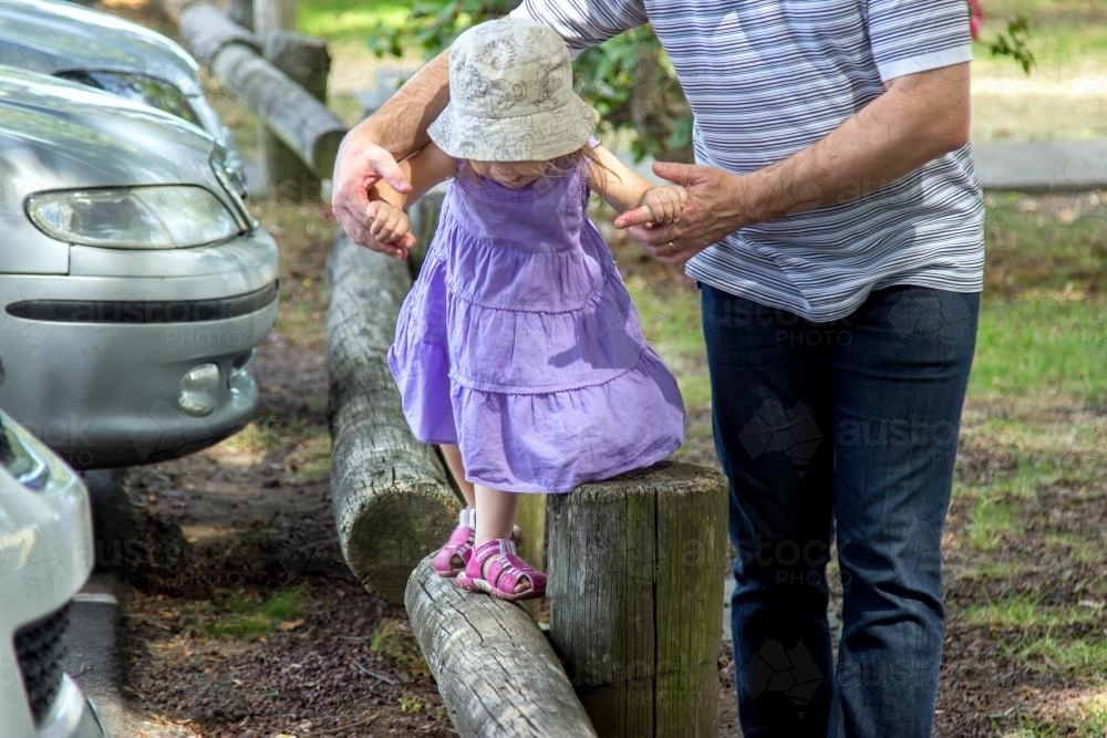 Father holding daughters hands as she walks along a pole at the park - Australian Stock Image