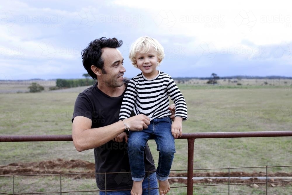 Father holding happy young son on an old farm gate on rural property - Australian Stock Image