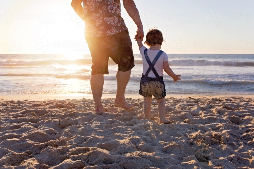 Father Holding His Daughter's Hand Walking Towards The Ocean At Sunset - Australian Stock Image