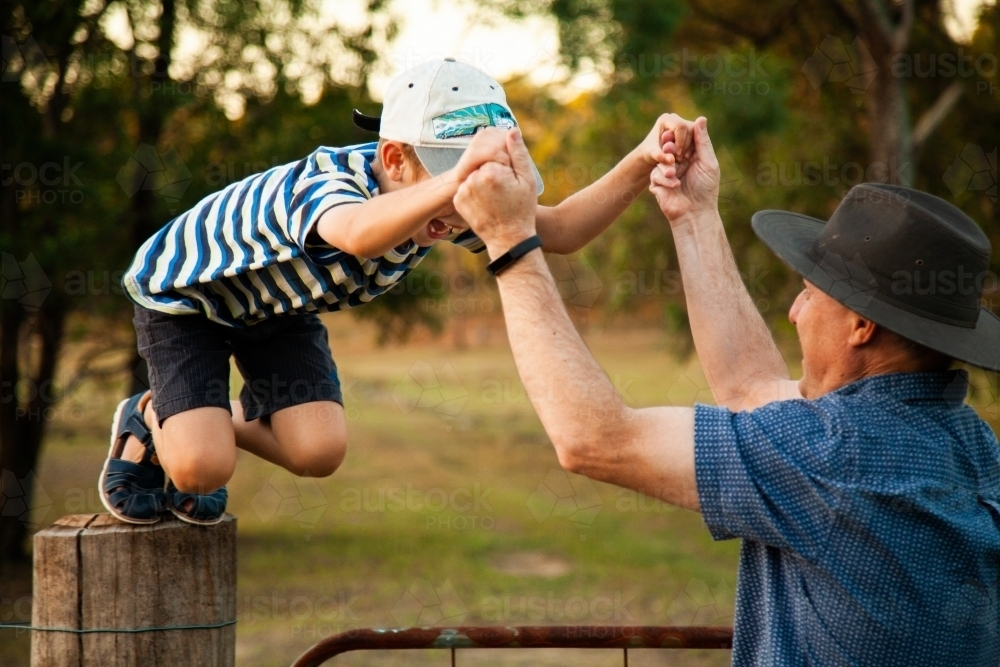 Father holding son's hands helping to jump off fence post - Australian Stock Image
