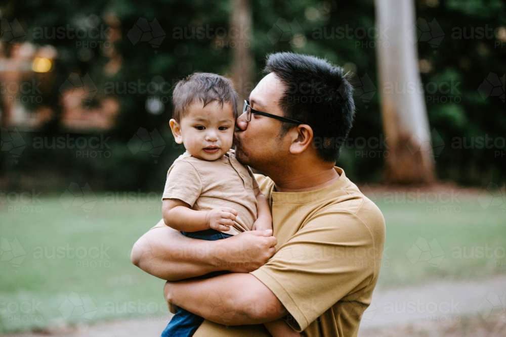 Father kissing baby boy cheek while holding him at the park - Australian Stock Image