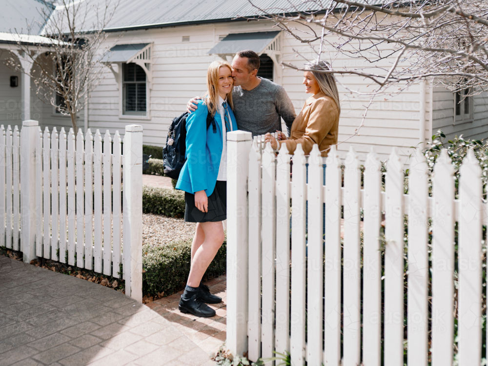 Father kissing teenage daughter leaving for school near picket fence with mum standing beside him - Australian Stock Image