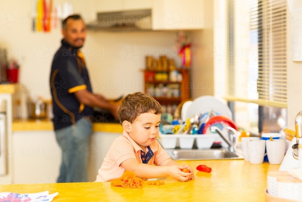 father looking on as son plays with dough in kitchen - Australian Stock Image