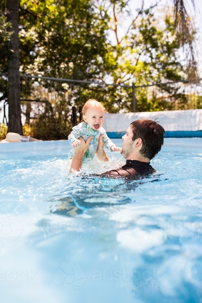 Father playing with baby in swimming pool dad lifting child high into air splashing in water - Australian Stock Image