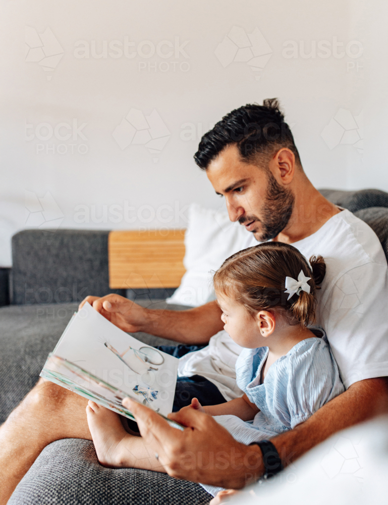 Father reading story with toddler daughter on the couch - Australian Stock Image