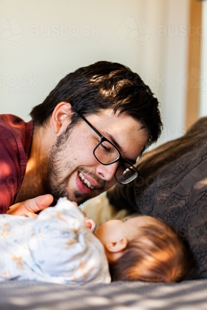 Father smiling and interacting with baby in morning sunlight on bed - Australian Stock Image
