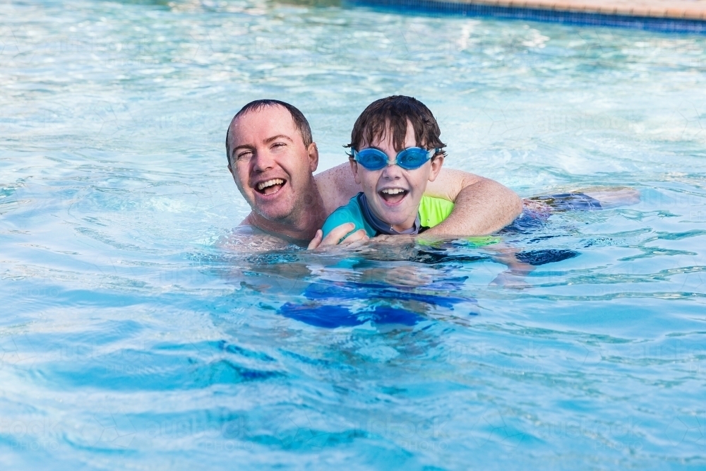 Father with arms around son wearing goggles playing in pool having fun laughing - Australian Stock Image