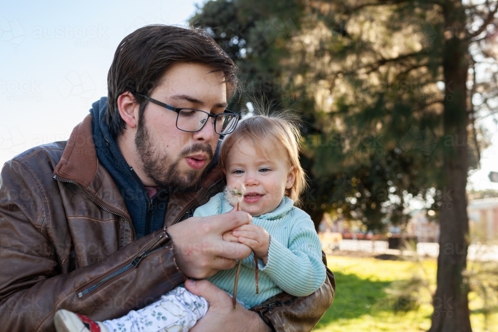Father with daughter at park hugging and blowing dandelion seed - Australian Stock Image