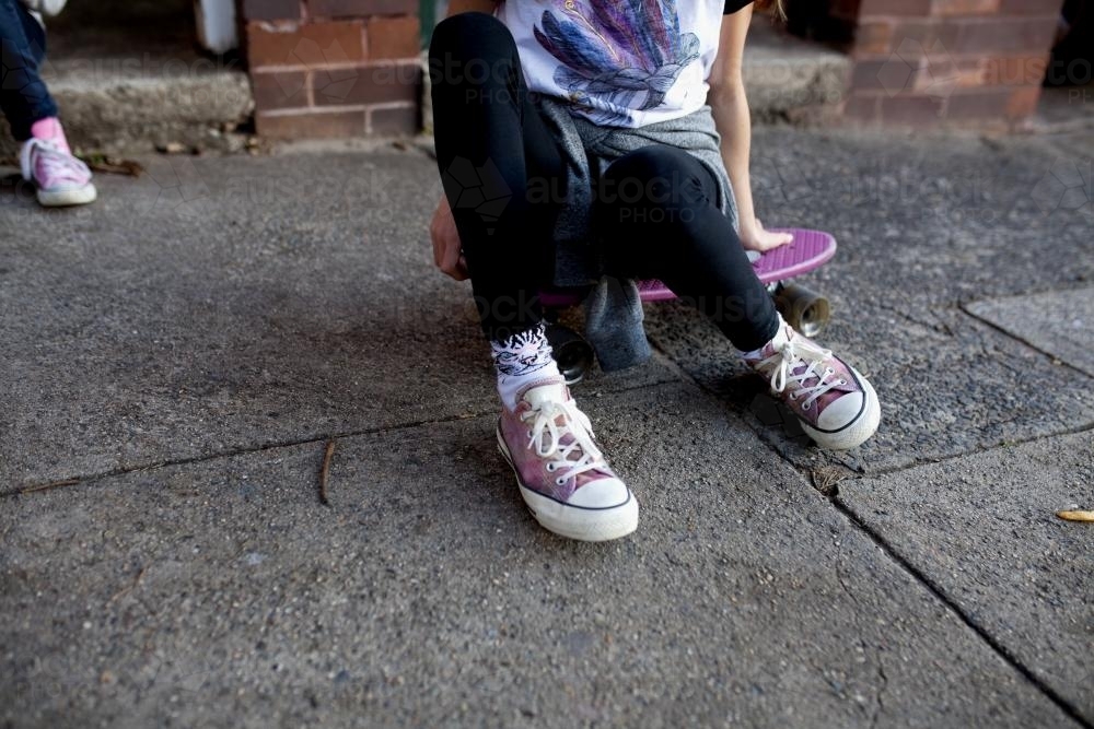 Feet and legs of a girl sitting on skateboard on footpath - Australian Stock Image