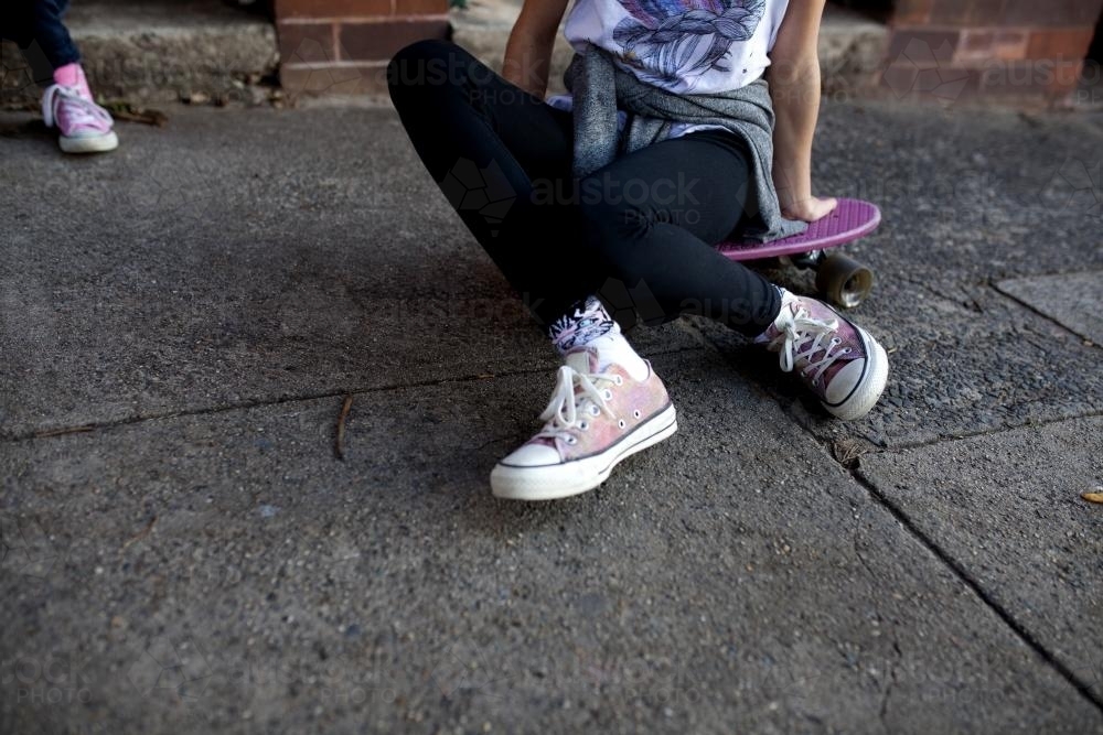 Feet and legs of a girl sitting on skateboard on footpath - Australian Stock Image