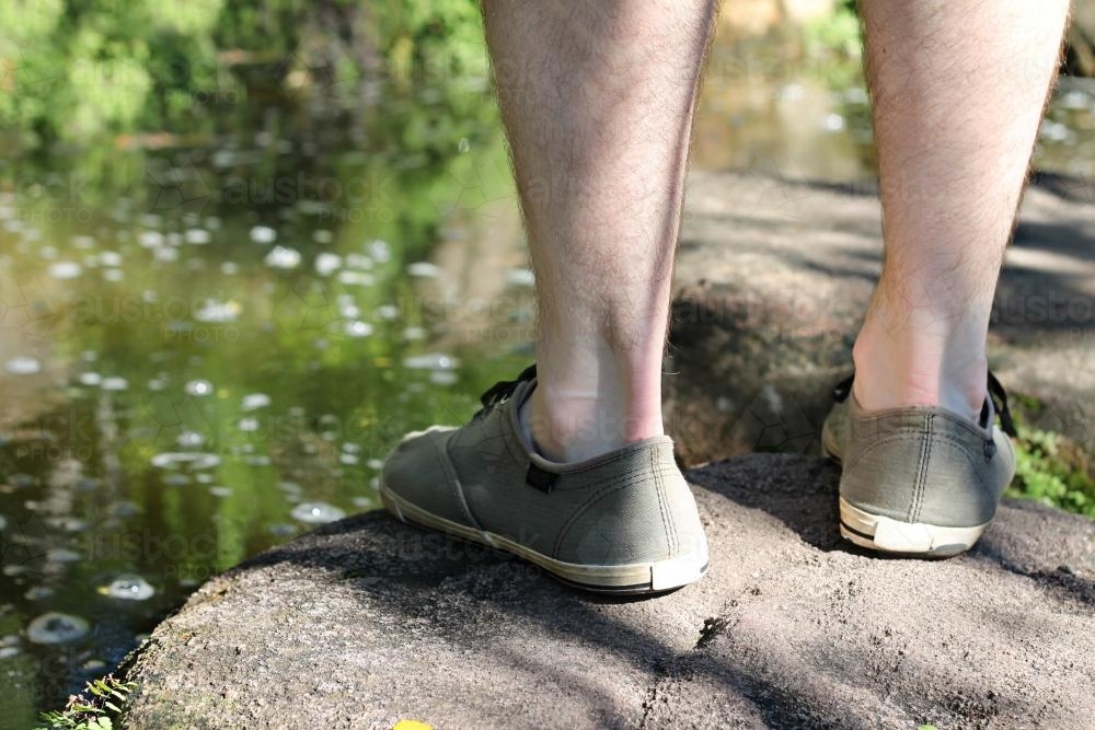 Feet of a man standing on stones beside water - Australian Stock Image
