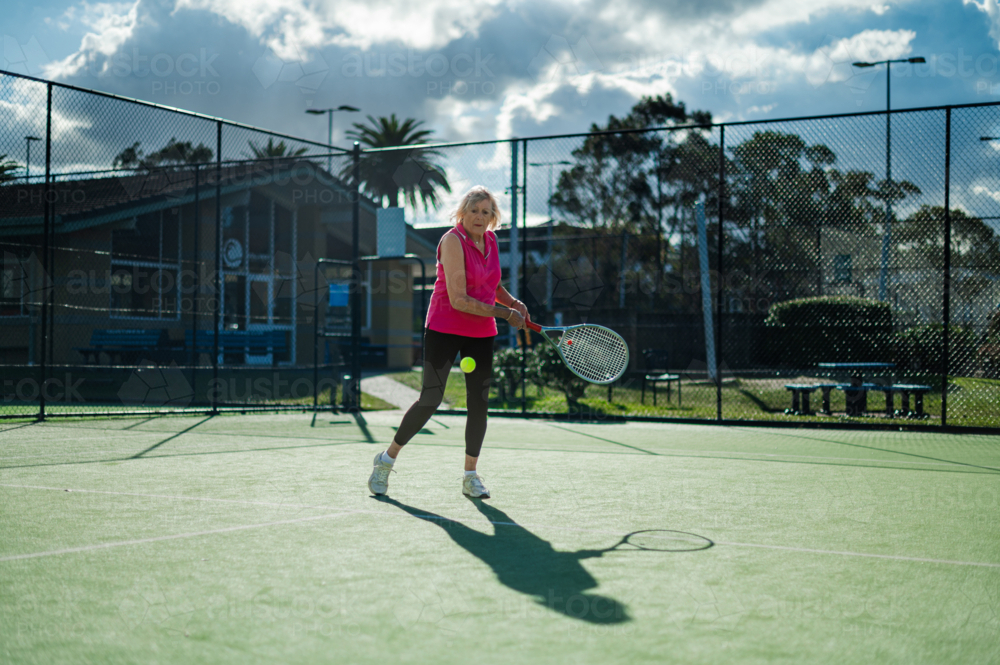 Female athlete hitting a tennis ball on a court surrounded by greenery and blue skies - Australian Stock Image