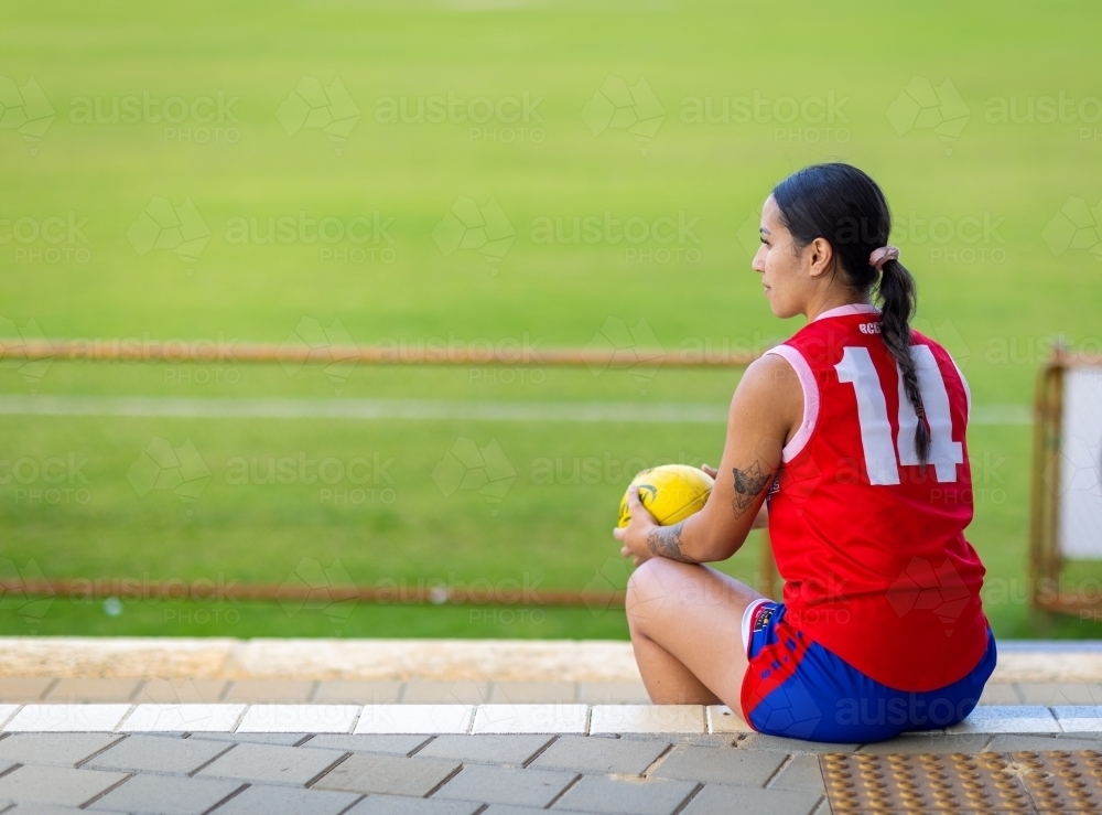 female aussie rules football player sitting on sidelines holding football - Australian Stock Image