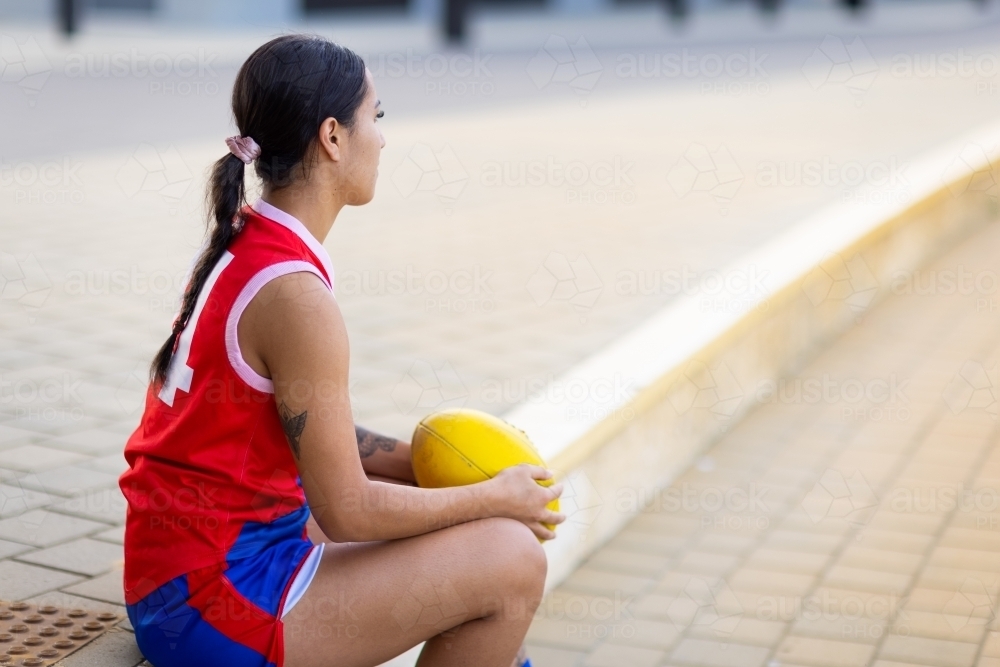 female aussie rules football player sitting on sidelines holding football - Australian Stock Image