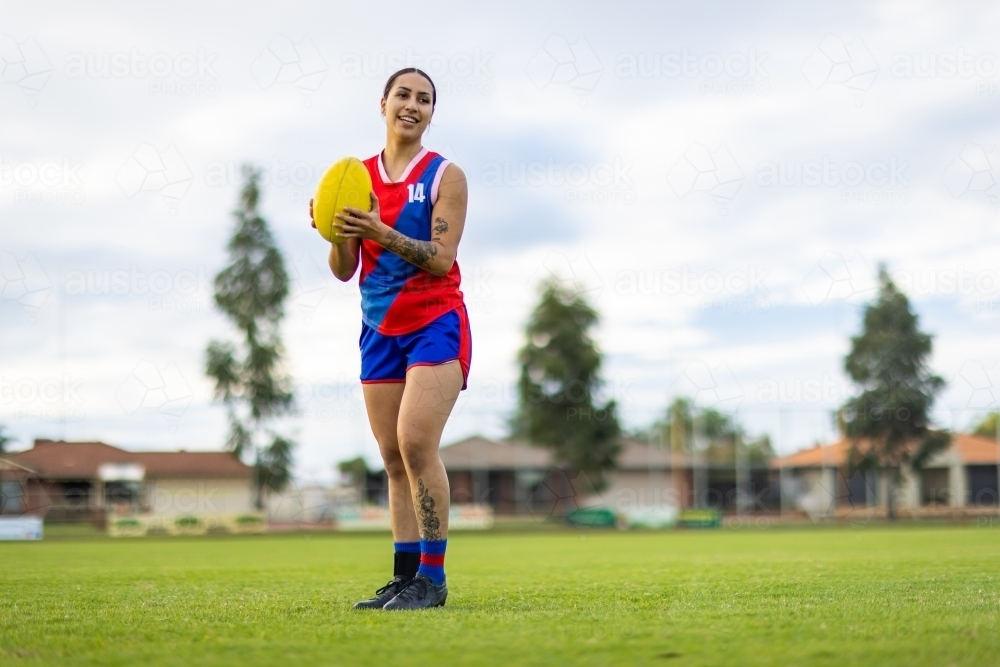 female aussie rules football player standing on footy oval with yellow leather football - Australian Stock Image