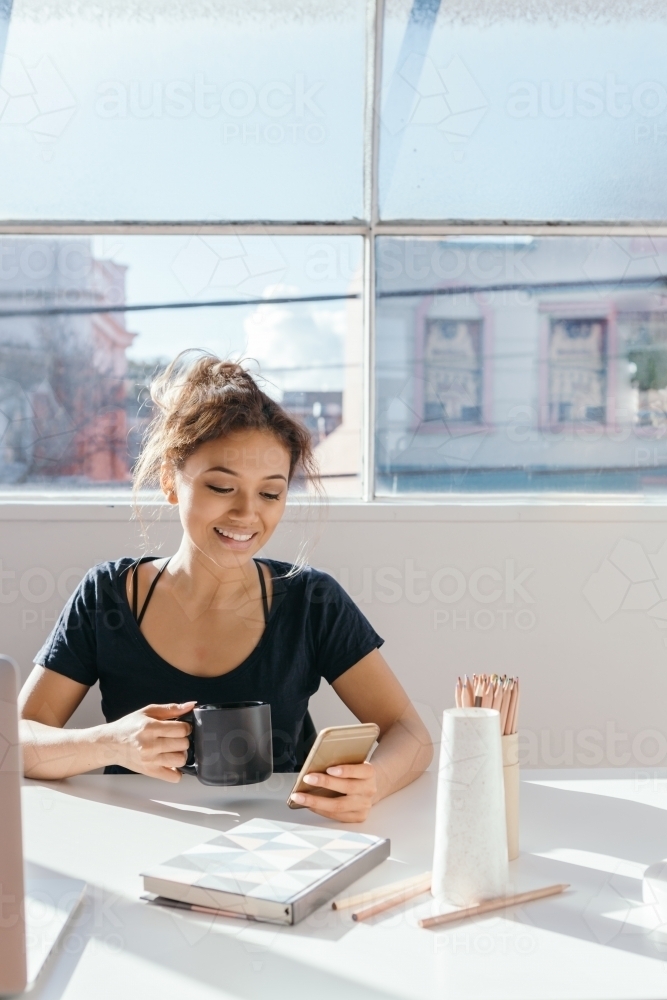 Female blogger checking her mobile device at a desk in a bright office - Australian Stock Image