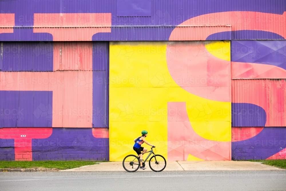 Female Cyclist Passing Bright Warehouse Wall - Australian Stock Image
