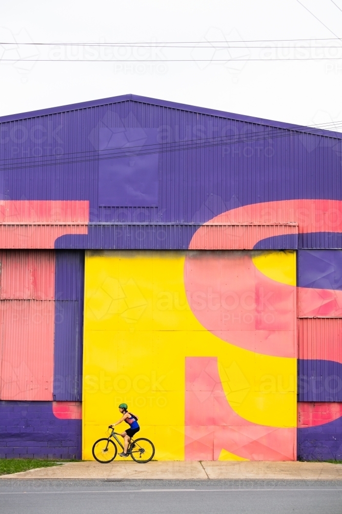 Female Cyclist Passing Bright Warehouse Wall - Australian Stock Image