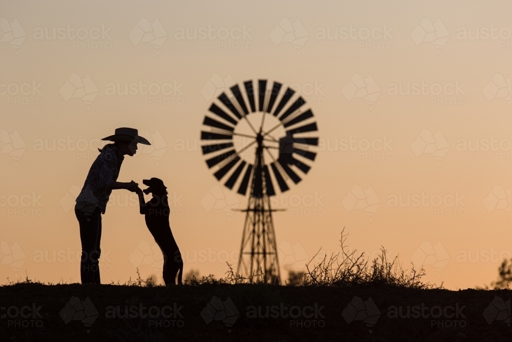Female farmer playing with dog in silhouette with windmill - Australian Stock Image