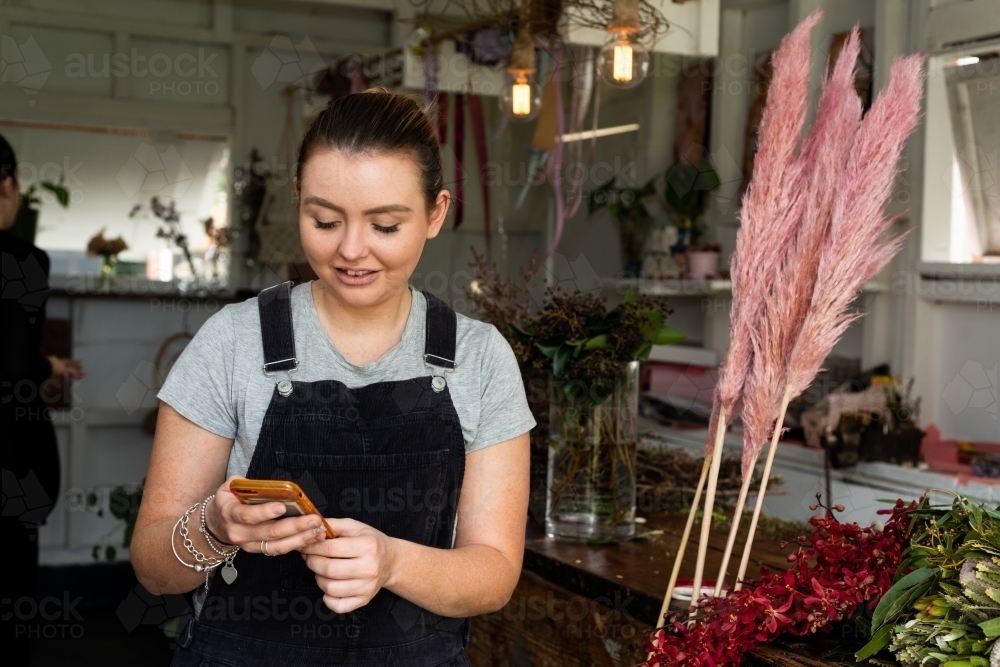 Female florist checking her mobile phone with pink Pampas Grass and darkened workshop behind - Australian Stock Image