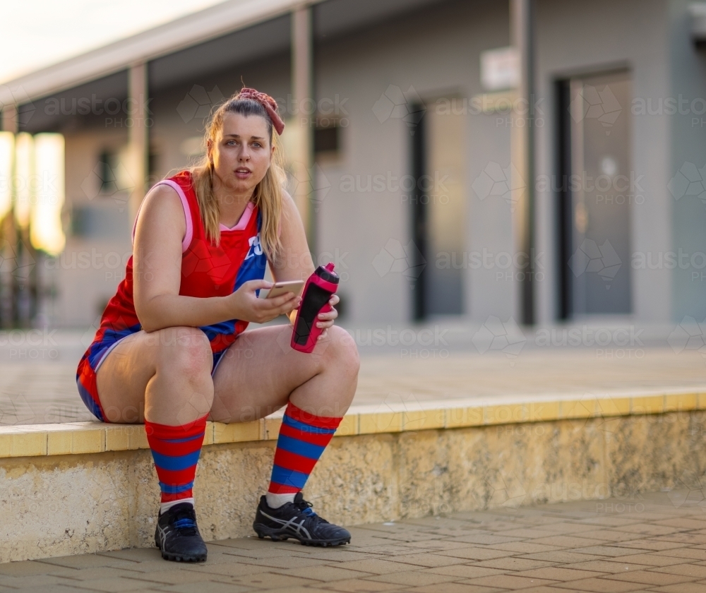 female football player seated on step with water bottle and phone - Australian Stock Image