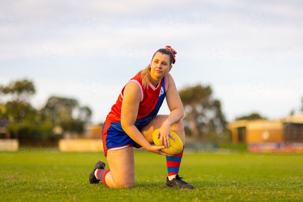 female footballer kneeling on grass on one knee holding football - Australian Stock Image