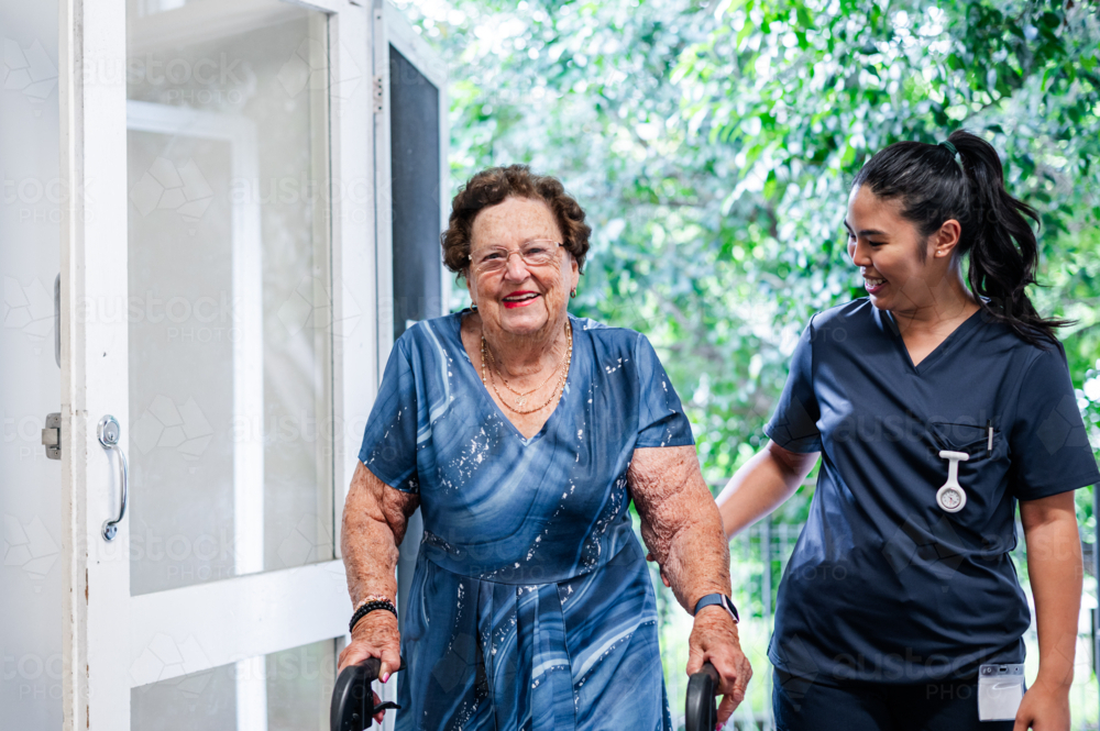 Female health caregiver supporting an elderly woman using a walker walk through the doorway - Australian Stock Image