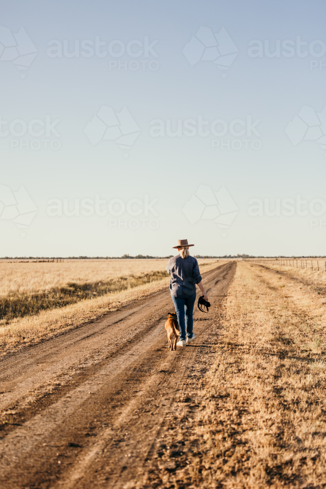 Female in distance walking along farm road carrying camera with dog - Australian Stock Image