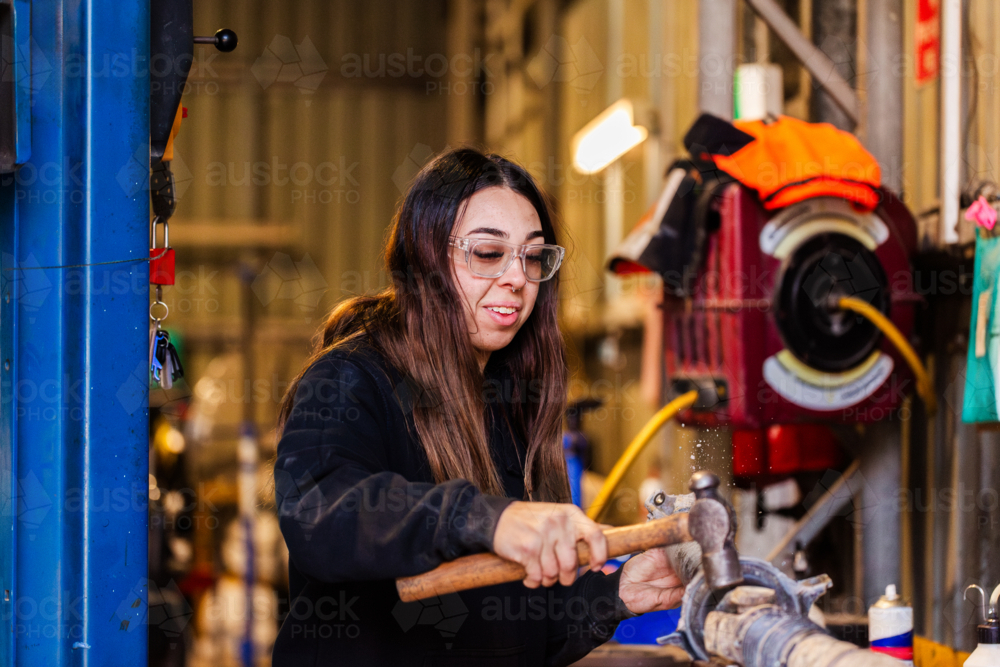 Female mechanic striking vehicle drive shaft with a hammer working on car repairs in workshop - Australian Stock Image