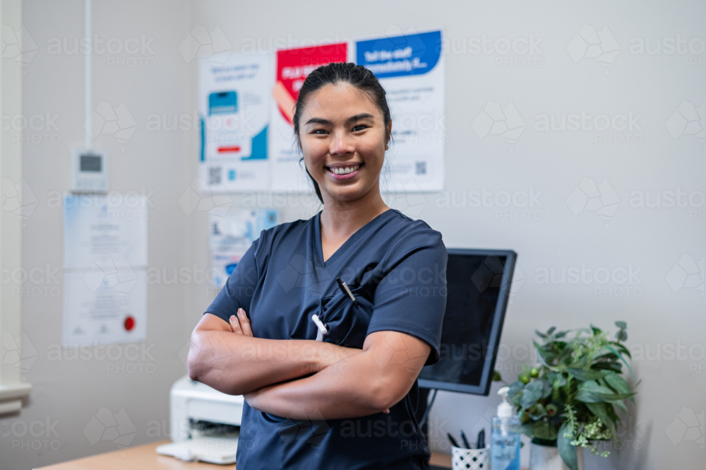 Female nurse in blue scrubs standing inside the clinic with her arms crossed - Australian Stock Image