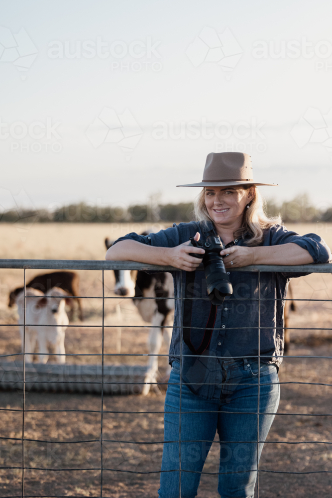 Female photographer holding camera leaning over a gate with cattle in background - Australian Stock Image
