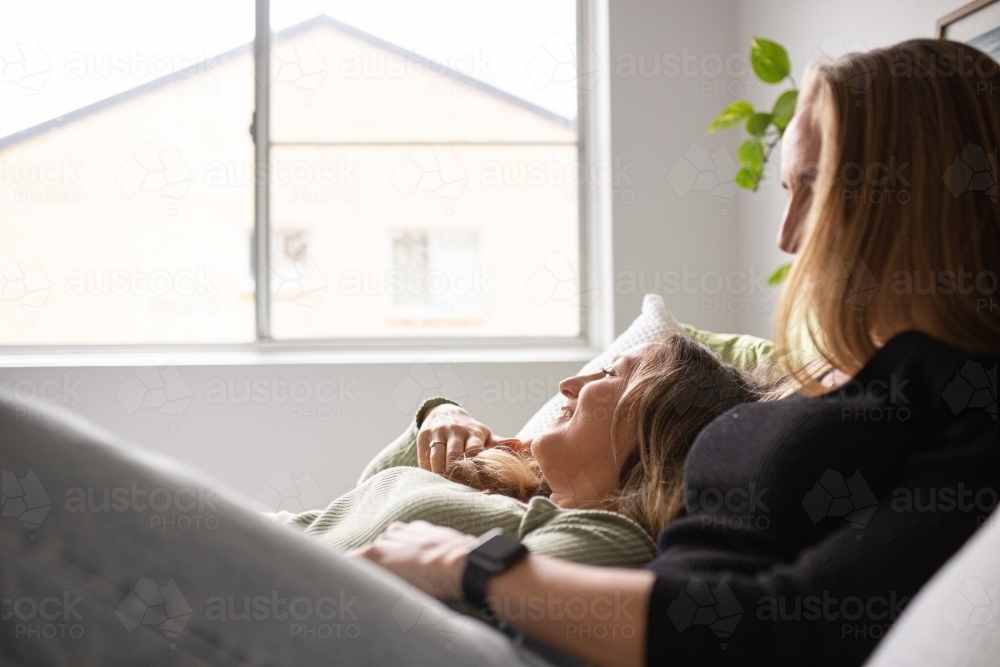 Female same sex couple  affectionately lying together on the bed, looking out the window - Australian Stock Image