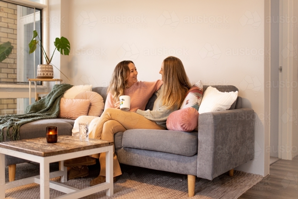 Female same sex couple  in the living room, talking and cuddling on the grey sofa - Australian Stock Image