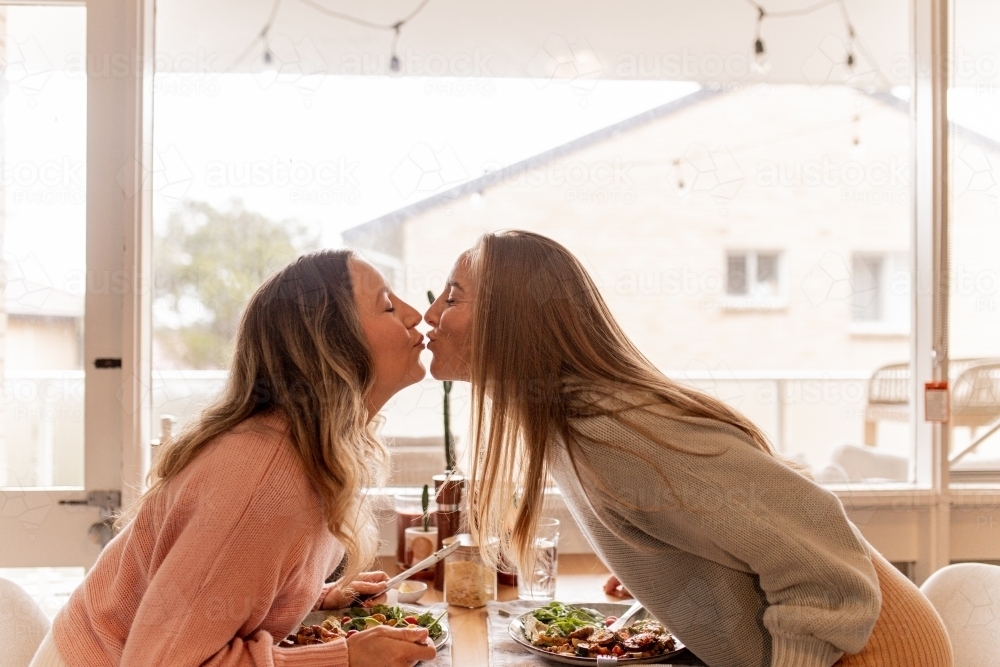 Female same sex couple kissing across the dining table at home - Australian Stock Image