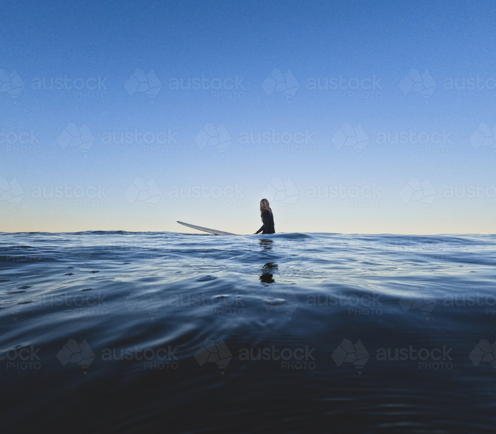 Female sitting on longboard surfboard looking out to see in glassy clear and still conditions - Australian Stock Image