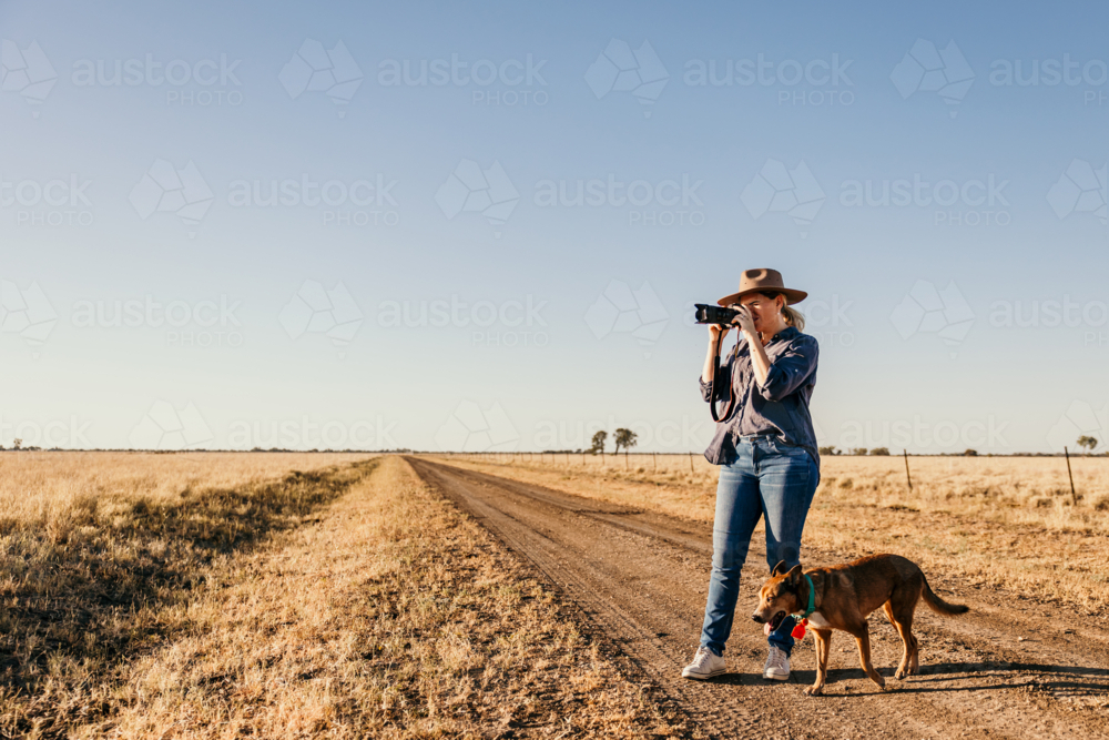 Female standing on road in paddock taking photo with dog - Australian Stock Image