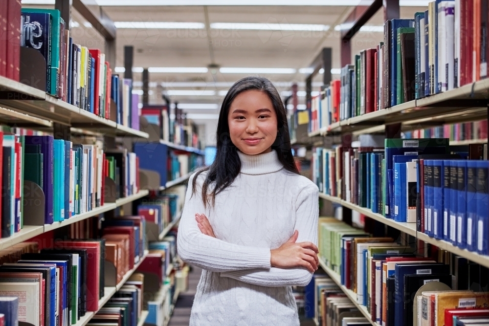 Female student standing with arms folded in library - Australian Stock Image
