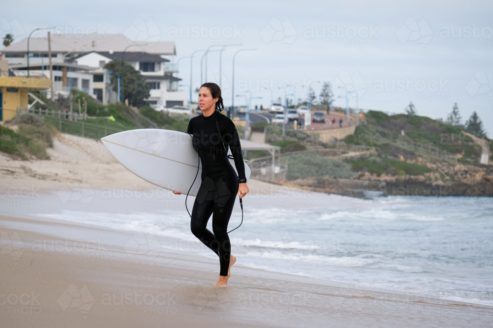 Female surfer in wetsuit walking out of ocean holding shortboard surfboard with coastal background - Australian Stock Image