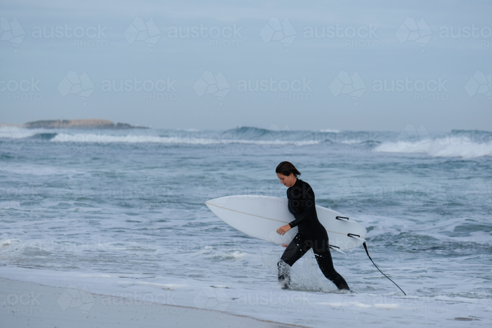Female surfer in wetsuit walking out of ocean holding shortboard surfboard with coastal background - Australian Stock Image