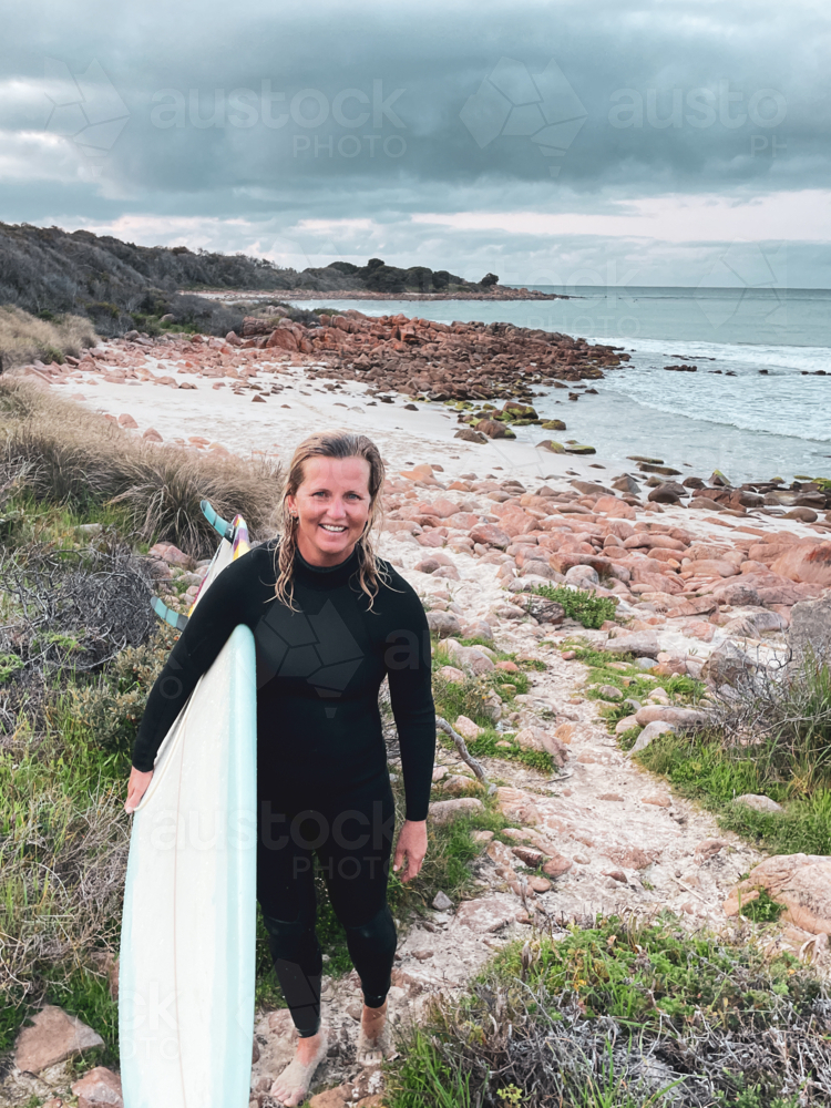 Female surfer walking with surfboard in wetsuit along sandy path with ocean coastal background - Australian Stock Image