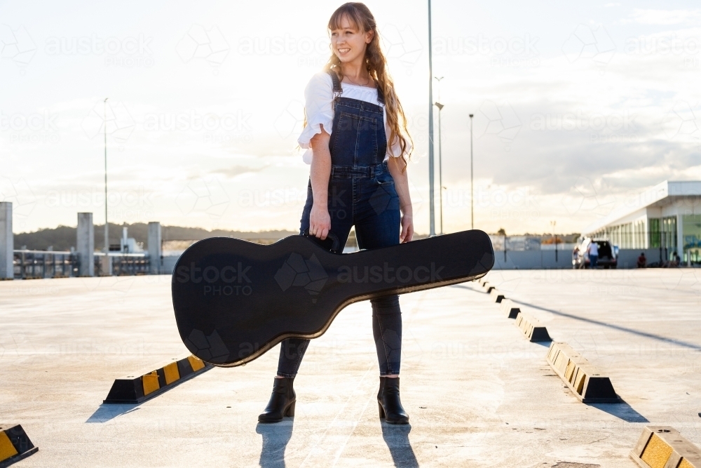 Female teenager posing in empty carpark with guitar case - Australian Stock Image