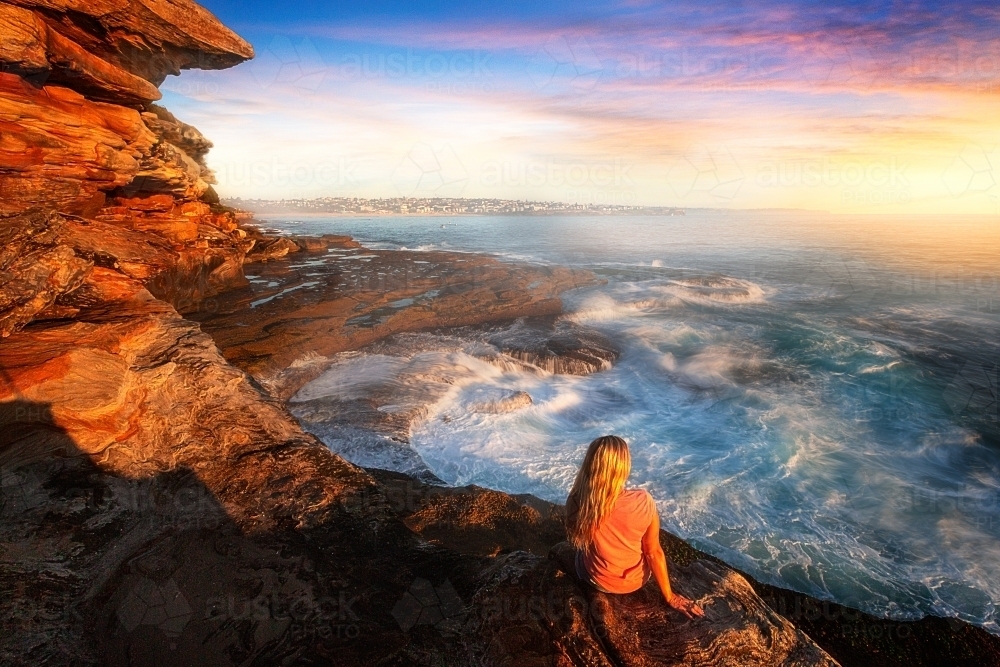 Female watches the ocean wash up and cascade like a waterfall and toss and swirl around ocean rocks - Australian Stock Image