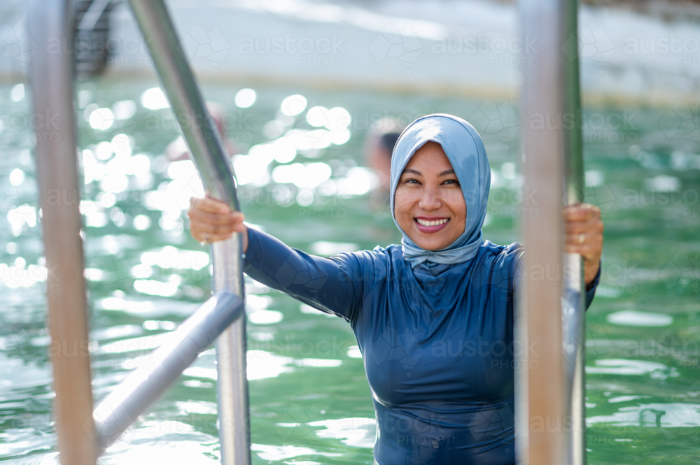 Filipina woman in a light blue headscarf holding on to metal handrail getting out of ocean pool - Australian Stock Image