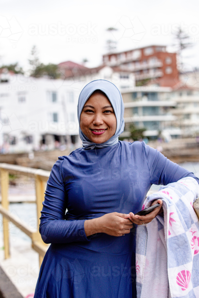 Filipina woman walking on coastal path holding a phone and a towel on the other hand - Australian Stock Image