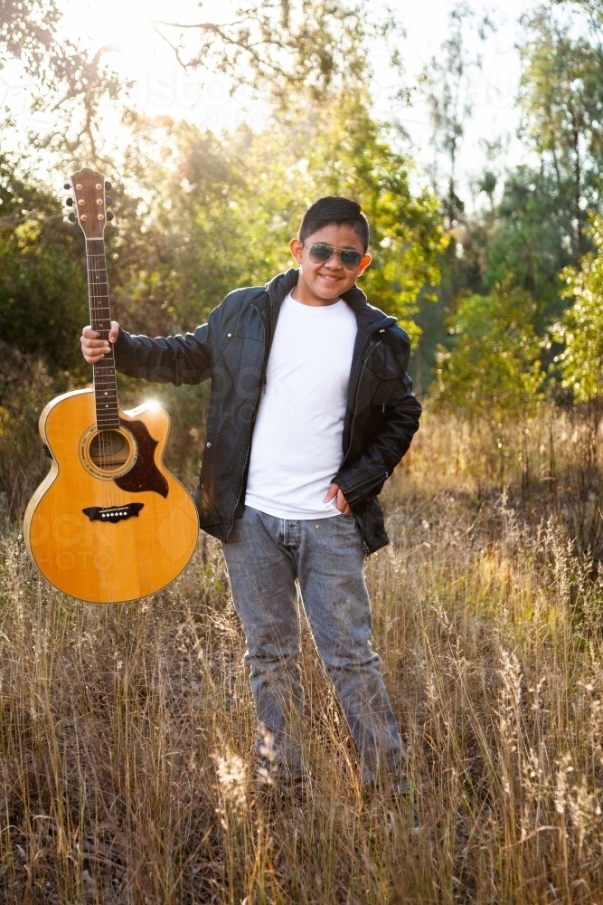 Filipino kid with guitar outside in bushland backlit with sun flare - Australian Stock Image