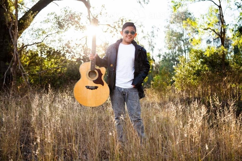 Filipino kid with guitar outside in bushland backlit with sun flare - Australian Stock Image