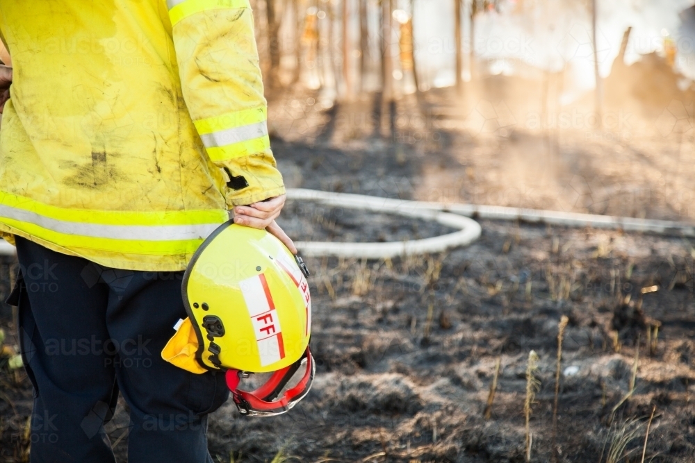 Fire fighter standing beside burnt grass holding safety helmet in hand - Australian Stock Image